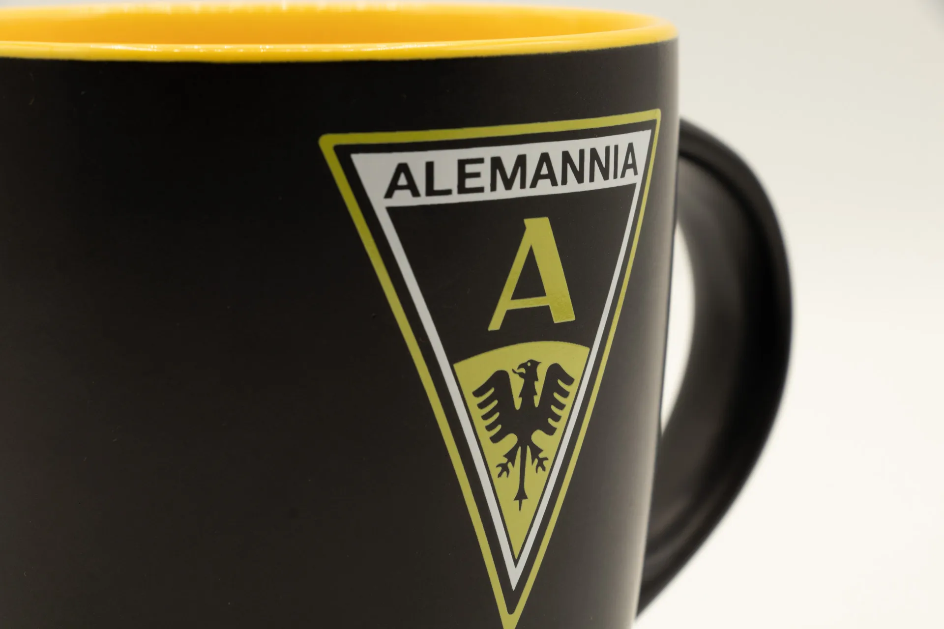 Schwarze matte Porzellantasse mit gelber Innenseite, mit einem Motiv zum Aufstiegslied „Aus meinem Traum bin ich heute aufgewacht…“ und dem Schriftzug „Alemannia“, 0,27 l